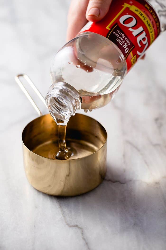 Hand pouring corn syrup into a brass measuring cup on a marble counter