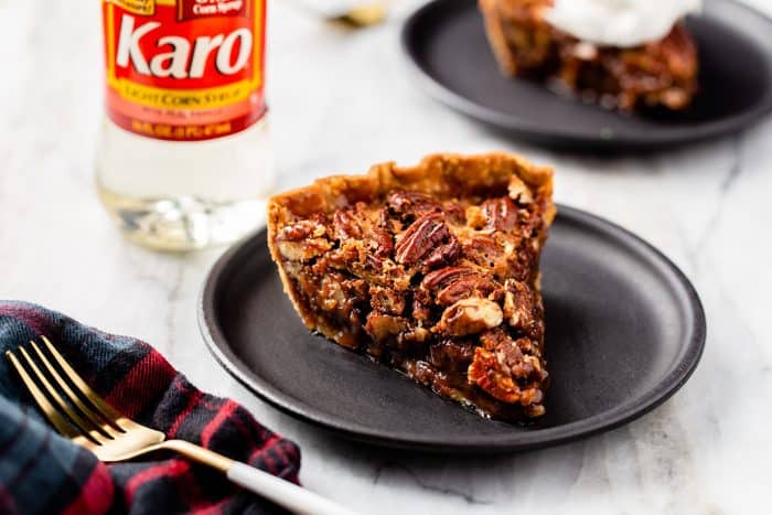 Slice of chocolate pecan pie on a black plate next to a bottle of corn syrup