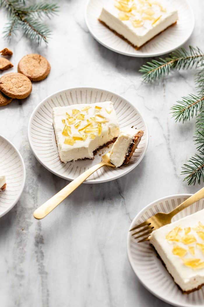 White plates holding slices of no-bake cheesecake bars arranged on a marble counter among sprigs of greenery