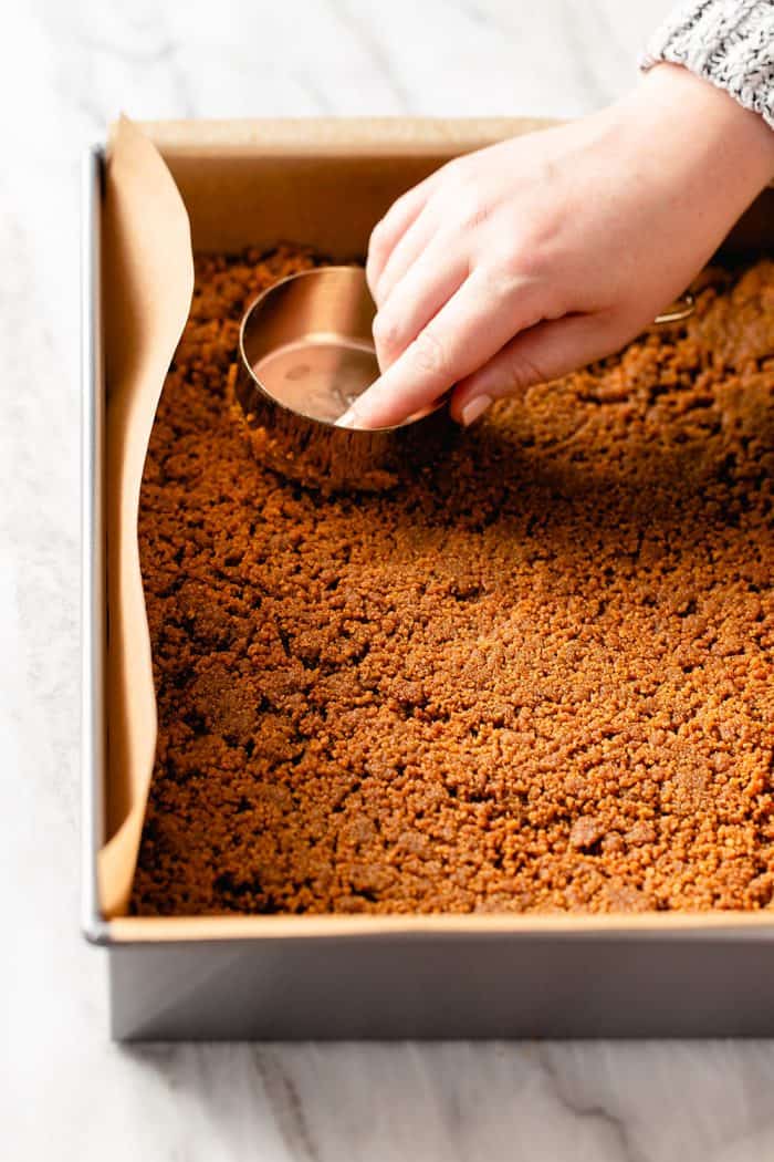 Hand using a measuring cup to press a cookie crust into a parchment-lined pan