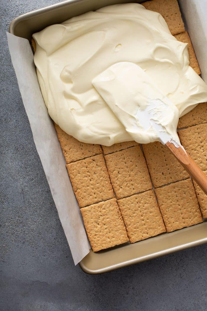 Spatula spreading the filling for banana cream pie eclair cake over a layer of graham crackers in a baking dish