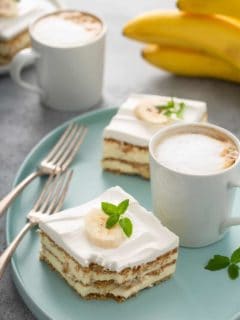 Two slices of banana eclair cake next to a latte and two forks on a blue platter, with another latte and a bunch of bananas in the background