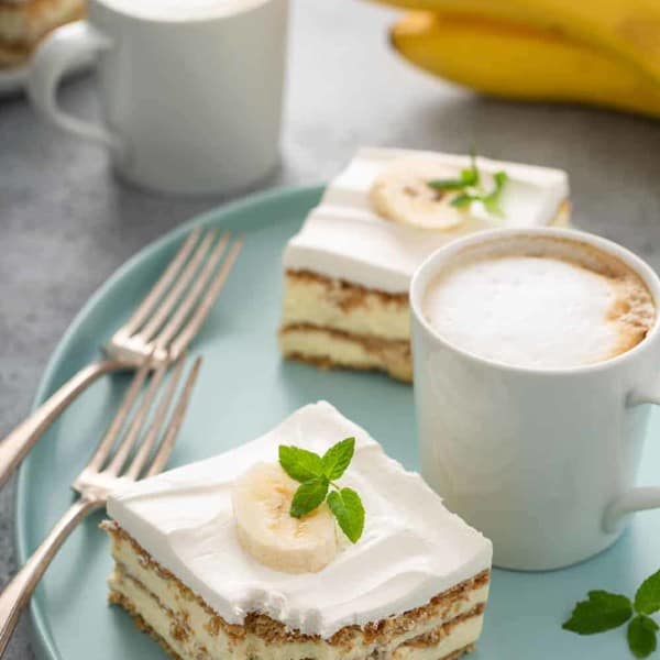 Two slices of banana eclair cake next to a latte and two forks on a blue platter, with another latte and a bunch of bananas in the background