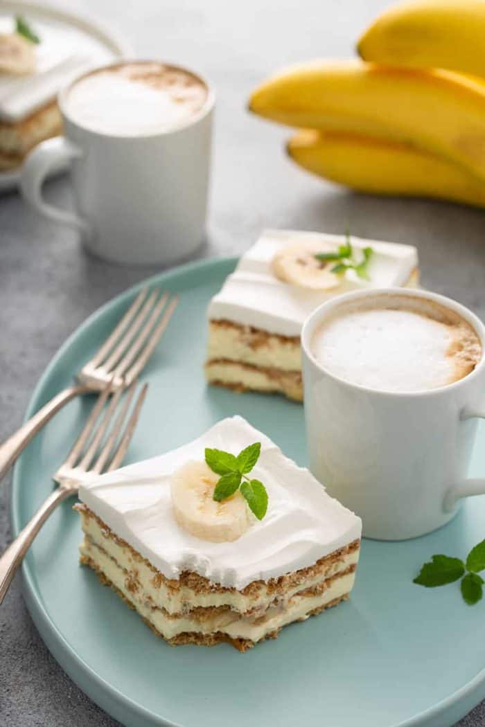 Two slices of banana eclair cake next to a latte and two forks on a blue platter, with another latte and a bunch of bananas in the background