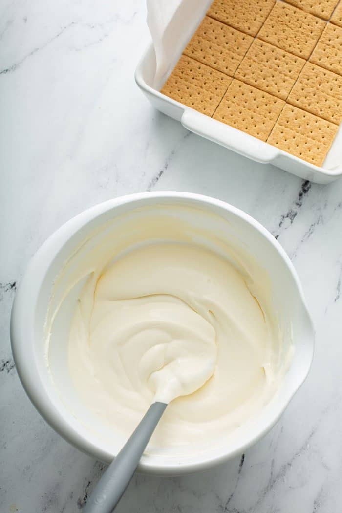 Bowl of cheesecake eclair cake filling next to a cake pan with a layer of graham crackers in the bottom