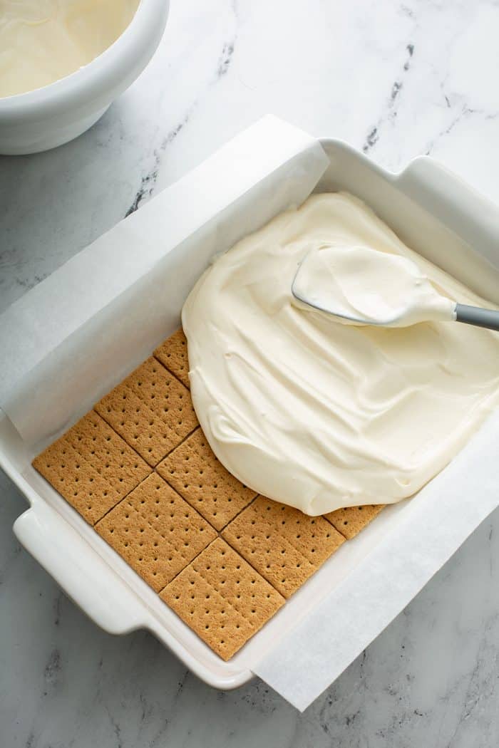 Spatula spreading the filling for no-bake cheesecake eclair cake on top of a layer of graham crackers in a white cake pan