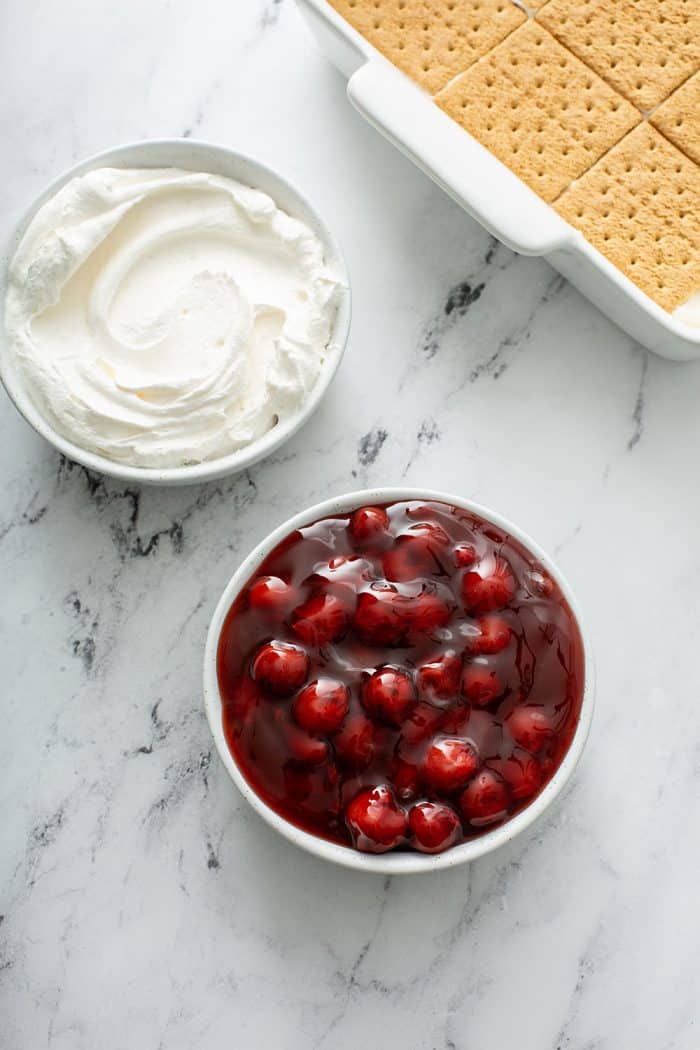 Cheesecake Eclair Cake in a white cake pan, bowl of whipped topping, and bowl of cherry pie filling on a marble countertop