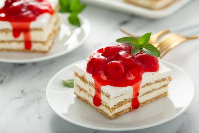 Slice of no-bake cheesecake eclair cake topped with cherry pie filling on a white plate