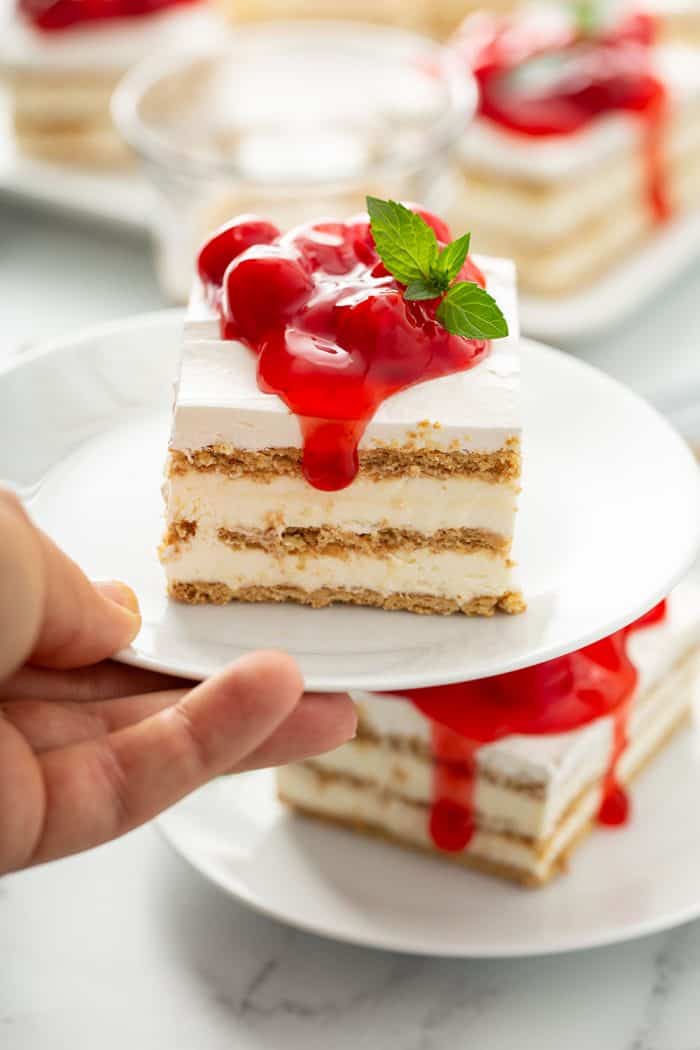 Hand holding up a plate with a slice of no-bake cheesecake eclair cake, showing layers of graham crackers and filling