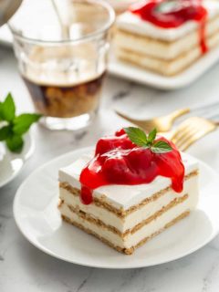 Slice of no-bake cheesecake eclair cake topped with cherry pie filling on a white plate, with a cup of espresso and more slices of eclair cake in the background