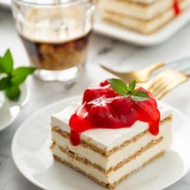 Slice of no-bake cheesecake eclair cake topped with cherry pie filling on a white plate, with a cup of espresso and more slices of eclair cake in the background