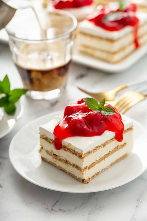 Slice of no-bake cheesecake eclair cake topped with cherry pie filling on a white plate, with a cup of espresso and more slices of eclair cake in the background