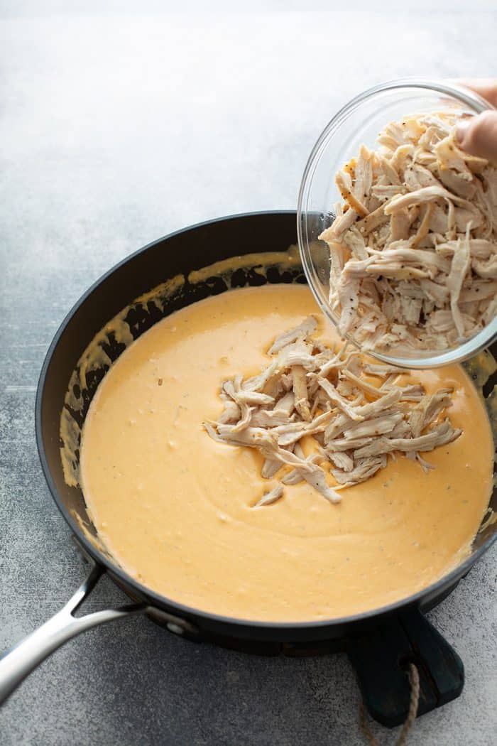 Shredded chicken being poured into a saucepan with the sauce for buffalo chicken dip