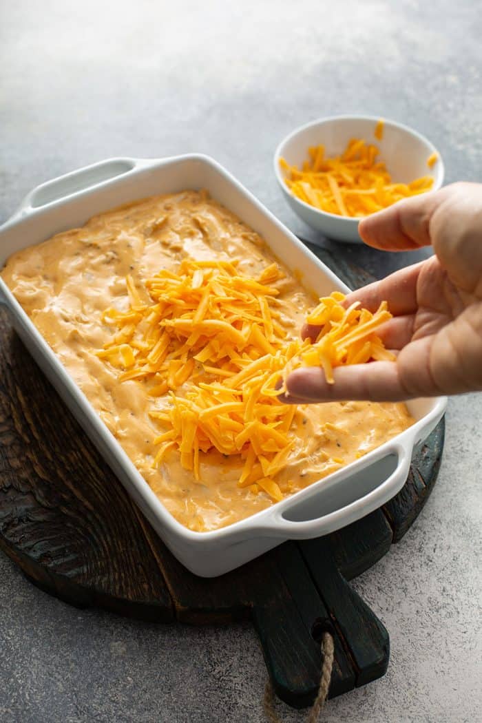 Hand sprinkling cheddar cheese on top of buffalo chicken dip in a white casserole dish