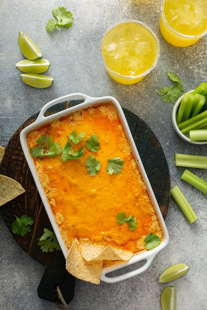 Overhead view of baked buffalo chicken dip in a white casserole dish surrounded by celery sticks and margaritas