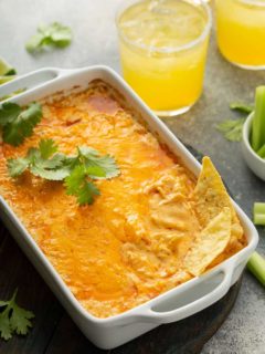 Tortillas sticking out of a buffalo chicken dip in a white casserole dish, with margaritas in the background