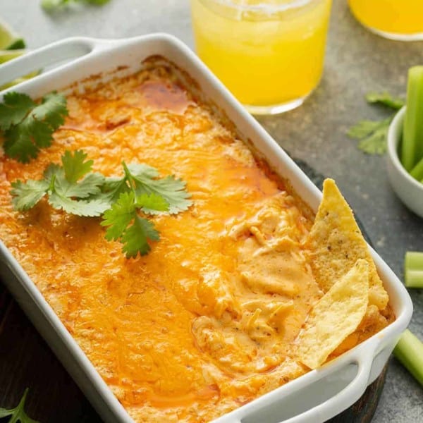 Tortillas sticking out of a buffalo chicken dip in a white casserole dish, with margaritas in the background