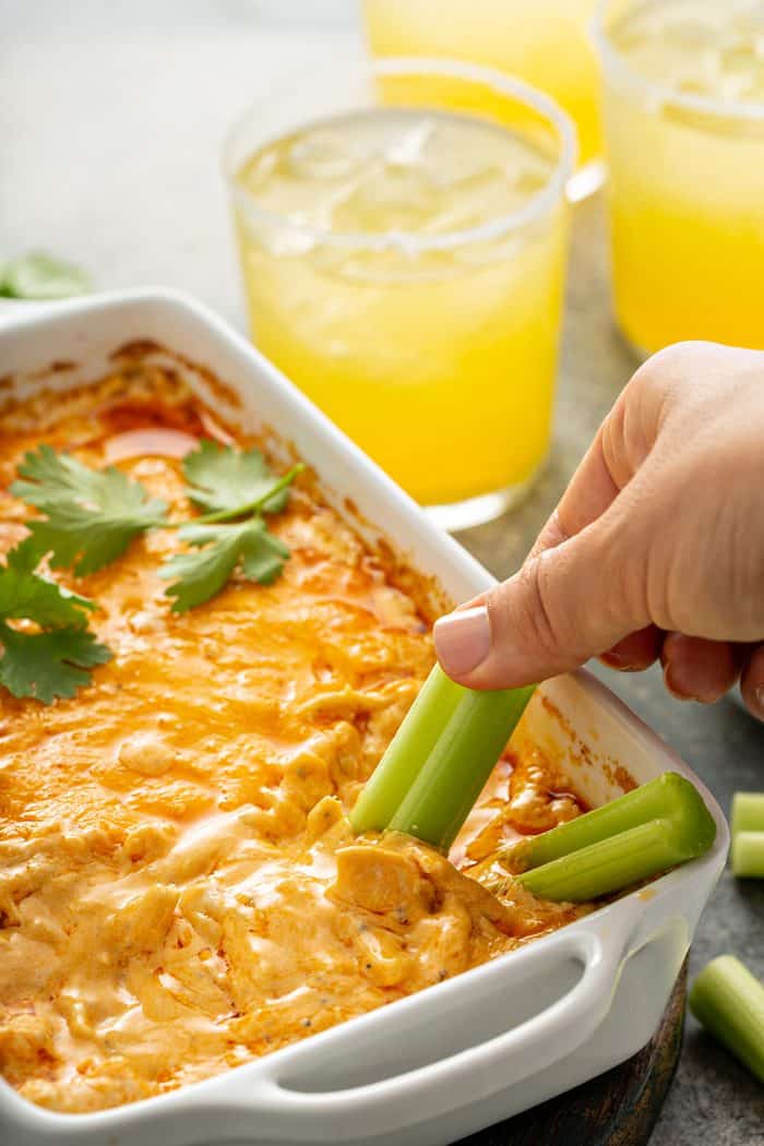 Hand dipping a celery stick into buffalo chicken dip in a white casserole dish