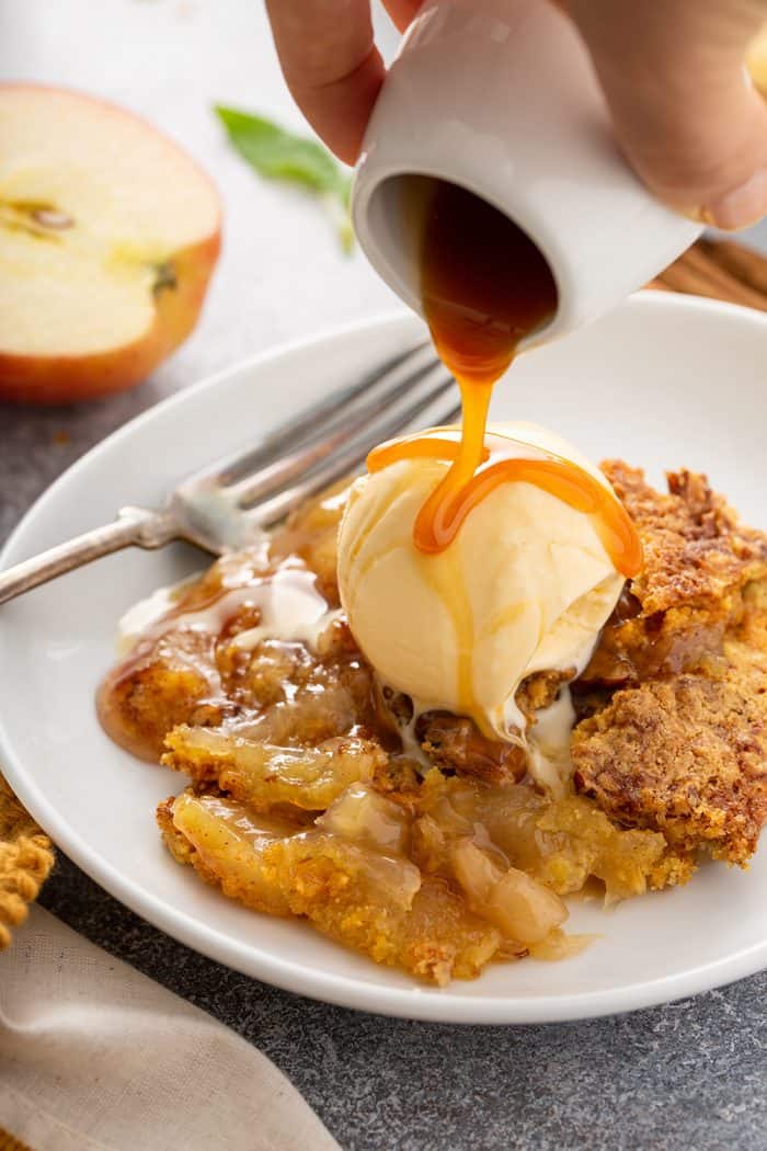 Hand pouring caramel sauce over plated apple dump cake topped with ice cream