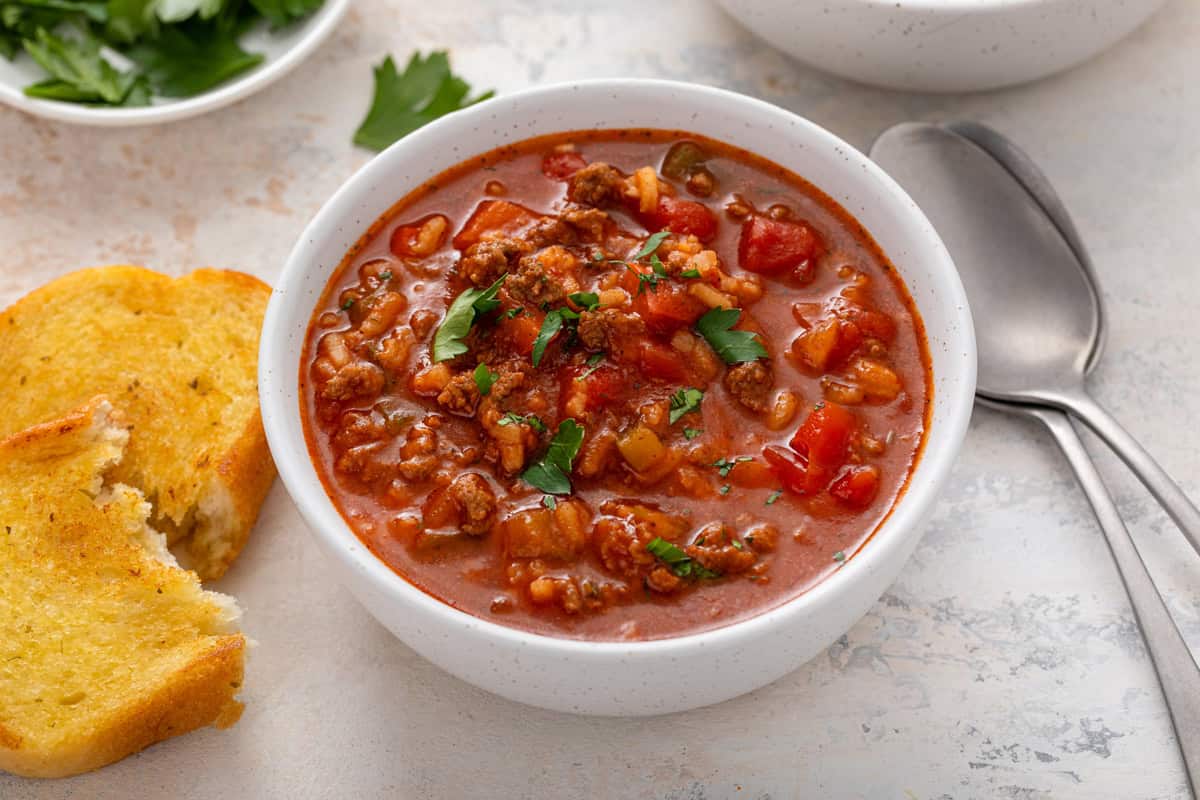 White bowl filled with stuffed pepper soup, garnished with parsley, resting next to texas toast on a countertop.