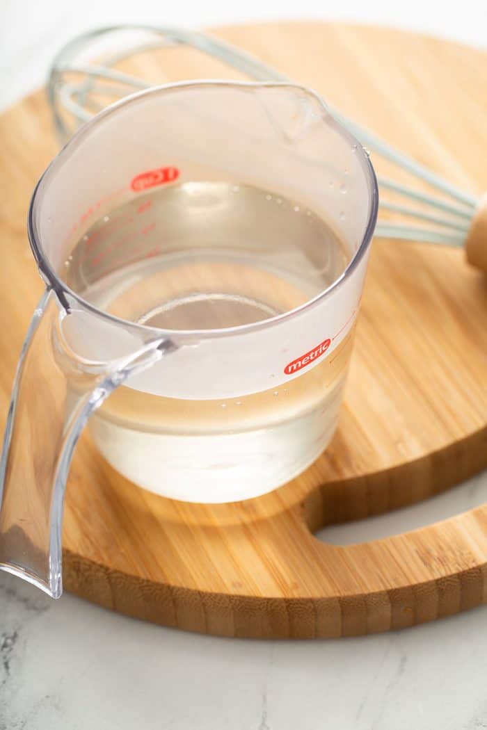 Measuring cup of simple syrup on a wooden board next to a whisk