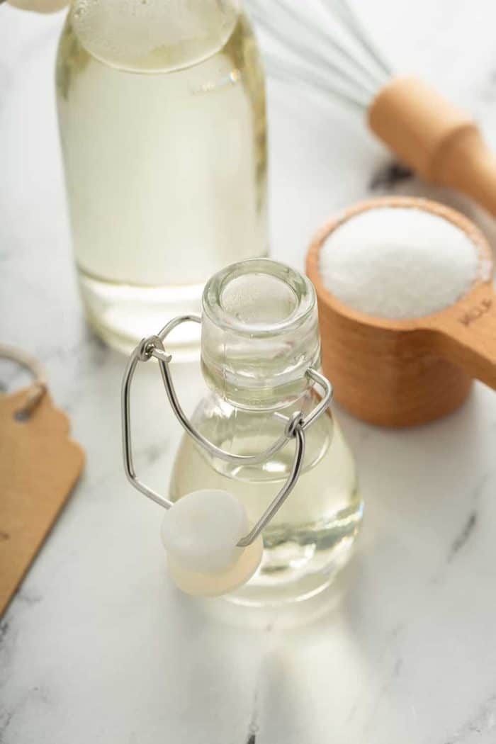 Small glass bottle of simple syrup on a marble counter with a measuring cup of sugar