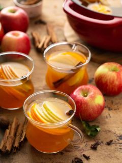 Three mugs of hot wassail garnished with sliced apples and cinnamon sticks with a dutch oven full of wassail in the background