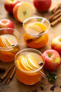 Three mugs of wassail garnished with sliced apples and cinnamon sticks, surrounded by more whole apples