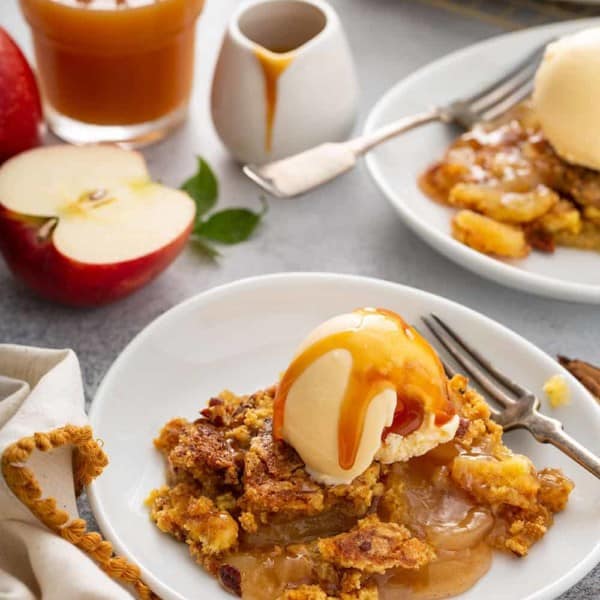 Two shallow bowls filled with apple dump cake topped with vanilla ice cream and caramel sauce