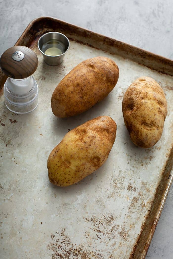 Three baking potatoes, salt and pepper on a sheet tray