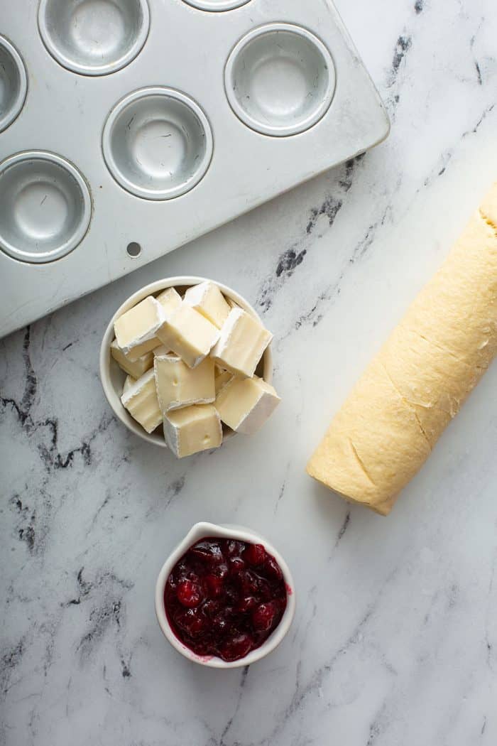 brie cheese, cranberry sauce, and crescent roll dough arranged on a marble counter next to a mini muffin tin