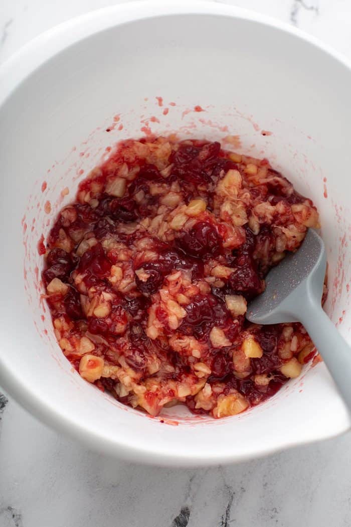 Spatula stirring together cranberry sauce and crushed pineapple in a white mixing bowl