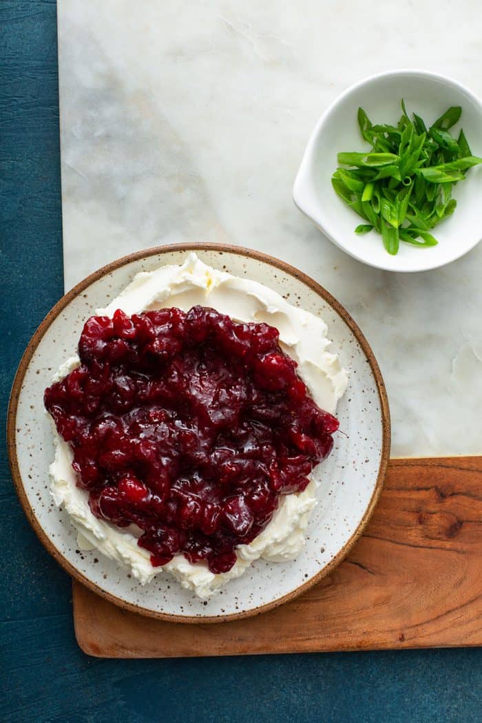 Cream cheese topped with cranberry sauce on a white plate next to a bowl of scallions