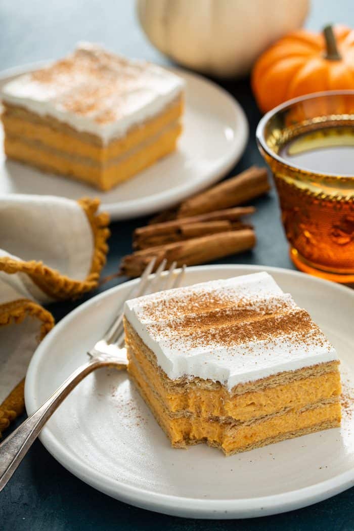 Slice of pumpkin eclair cake with a bite taken out of the corner on a white plate. A second piece of eclair cake is in the background