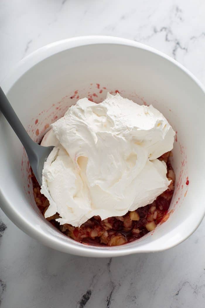 Spatula adding whipped topping to cranberry sauce and pineapple in a white mixing bowl