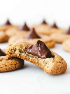Peanut Butter Blossom cookies made with peanut butter cookie dough and a Hershey's Chocolate Kiss. One cookie has a bite taken out of it, and is gently leaning against another. There are more cookies in the background.