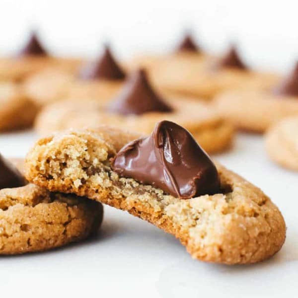 Peanut Butter Blossom cookies made with peanut butter cookie dough and a Hershey's Chocolate Kiss. One cookie has a bite taken out of it, and is gently leaning against another. There are more cookies in the background.