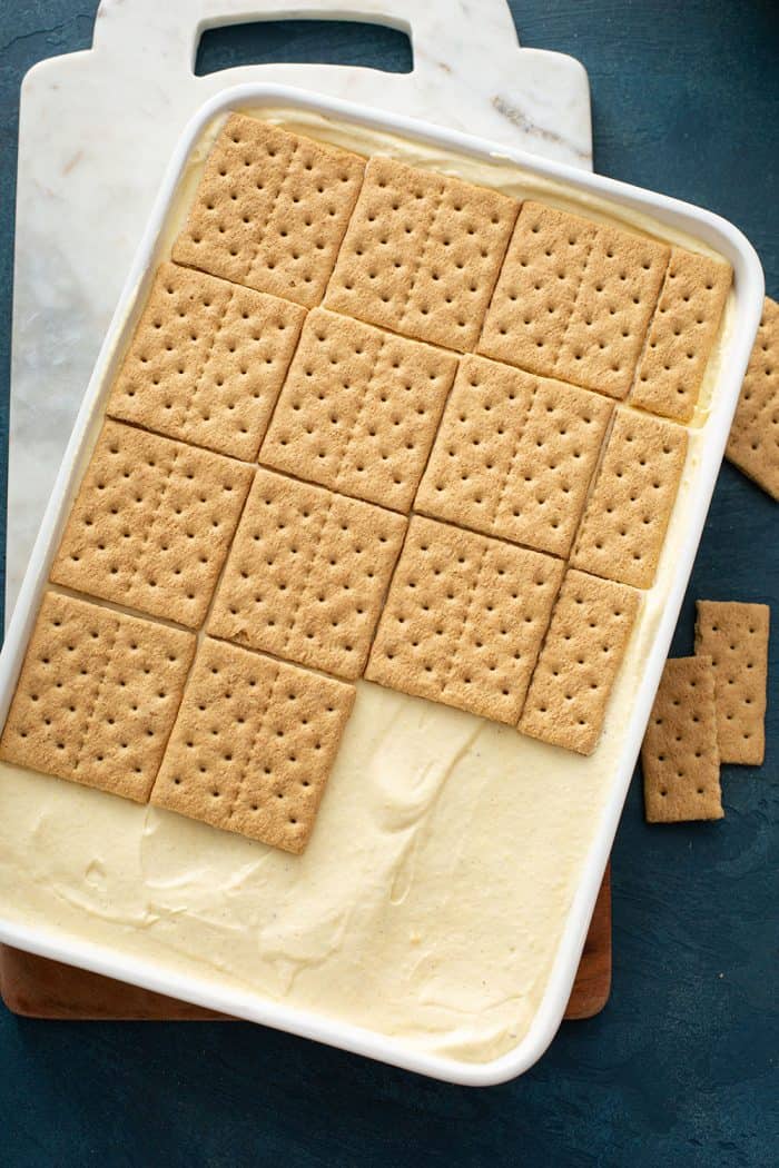 Cinnamon graham crackers being arranged on top of a pudding layer of an eclair cake