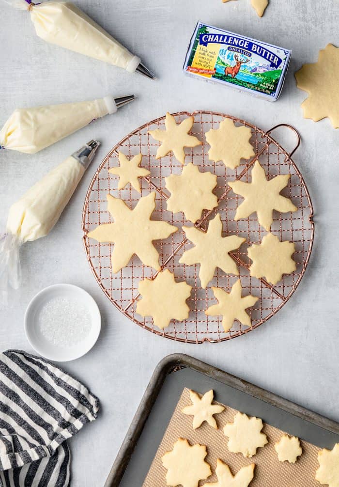 Baked sugar cookies shaped like snowflakes on a wire rack next to piping bags full of white buttercream frosting