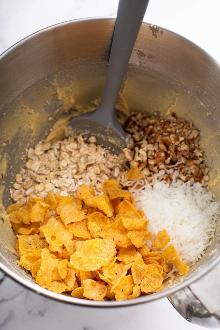 Spatula stirring oats, pecans, coconut, and cornflakes into cookie dough in a metal mixing bowl