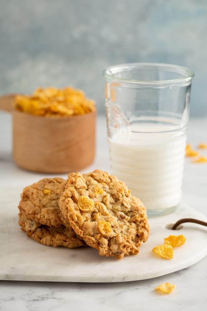 Three cornflake cookies next to a glass of milk with a measuring cup of cornflakes in the background