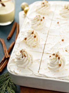 Close up of sliced eggnog eclair cake in a white cake pan next to cinnamon sticks and a glass of eggnog