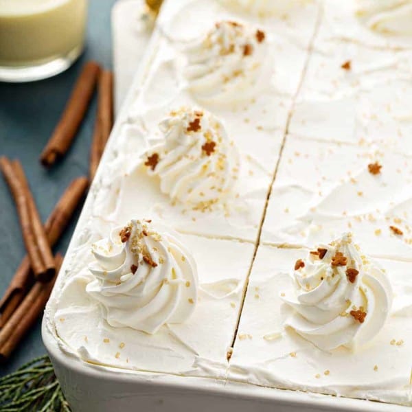 Close up of sliced eggnog eclair cake in a white cake pan next to cinnamon sticks and a glass of eggnog