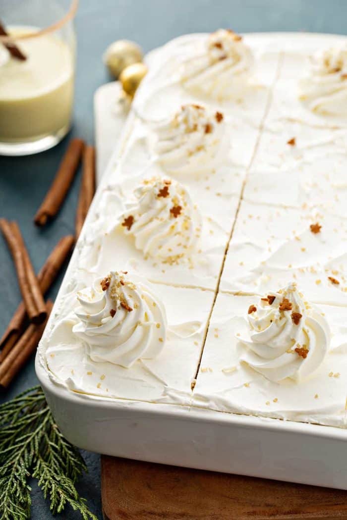 Close up of sliced eggnog eclair cake in a white cake pan next to cinnamon sticks and a glass of eggnog