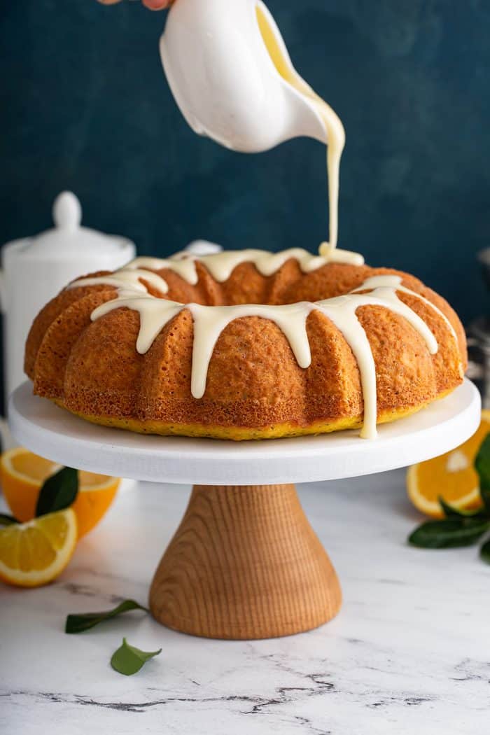 Glaze being poured over a citrus poppy seed cake on a white cake stand