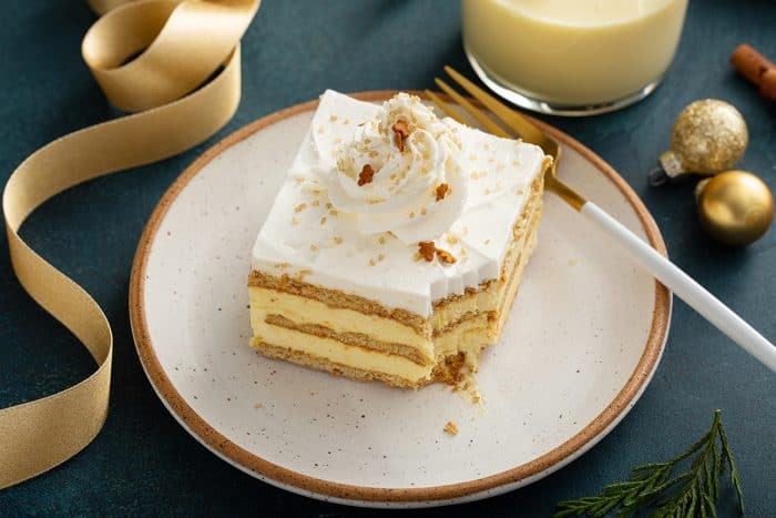 Slice of eggnog eclair cake with a bite taken out of it on a white plate next to a glass of eggnog