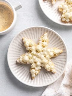 Snowflake sugar cookie on a white plate next to a cup of coffee