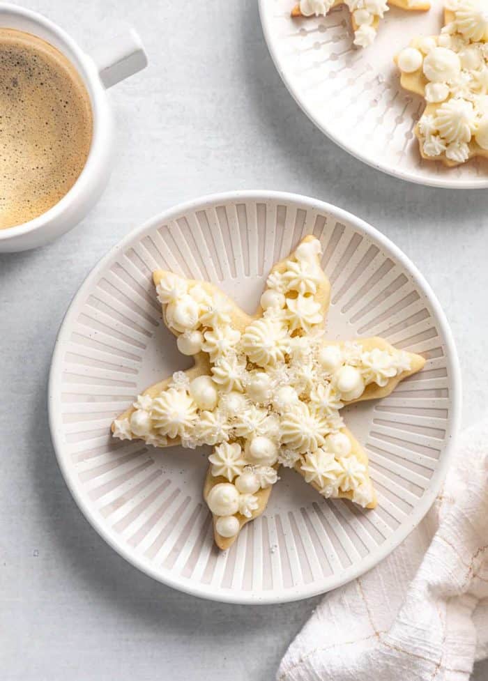 Snowflake sugar cookie on a white plate next to a cup of coffee