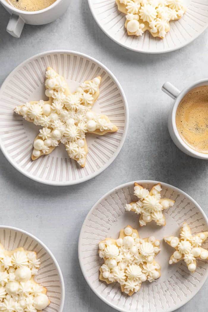 Assorted snowflake sugar cookies on white plates next to cups of coffee