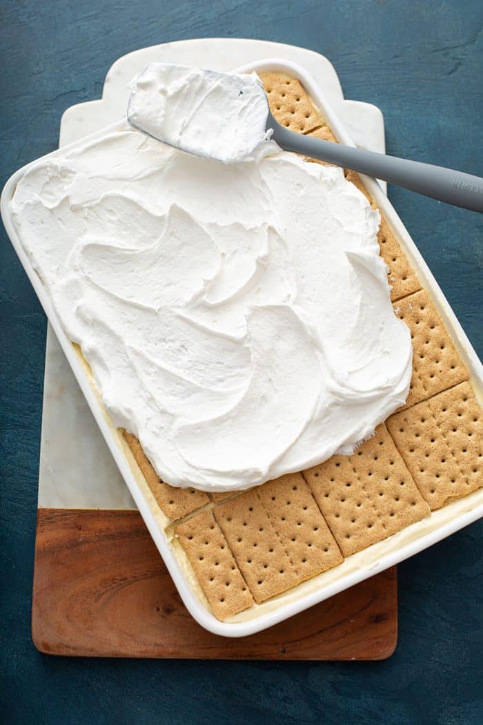 Whipped topping being spread on top of an eggnog eclair cake in a white cake pan